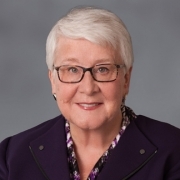 A professional portrait of attorney, Susan Coler. She has cropped white hair and wears a professional purple suit. 