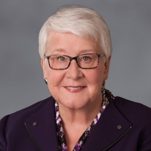 A closely cropped portrait of Susan Coler, attorney. White-haired and smiling in a smart purple suit, she's seated in front of a professional gray background.