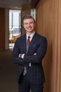 A portrait of young attorney, Colin. Confidently standing with crossed arms in a professional plaid suit and stands in an office and smiles at the camera.