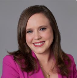 Professional portrait of attorney, Emma Denny. She wears a bright smile, pink suit and sits in front of a gray portrait studio background. 