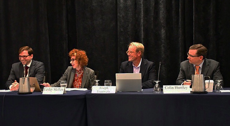 A panel of four professionals, three men and one woman, sit in front of a black velvet curtain. They smile as the woman presents from her laptop. 