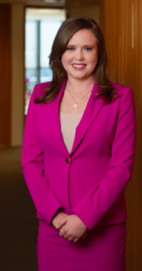 Attorney, Emma Denny stands, smiling for a portrait in a professional pink suit in a office with natural lighting. Her brown hair is neatly styled, falling over her shoulders. 