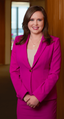 Standing in a neat office, attorney Emma Denny smiles for a portrait. Wearing a bright smile and pink suit, the brown-haired woman looks confidently into the camera. 
