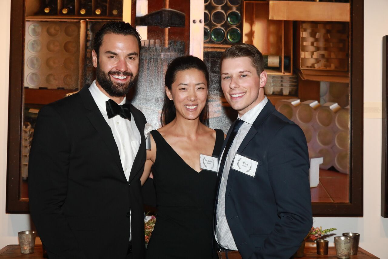 Two men and a woman stand in front of a modern photograph. The two young men wear suits and one wears a name tag. The woman wears a black dress and a name tag reading "Dr. Judith."