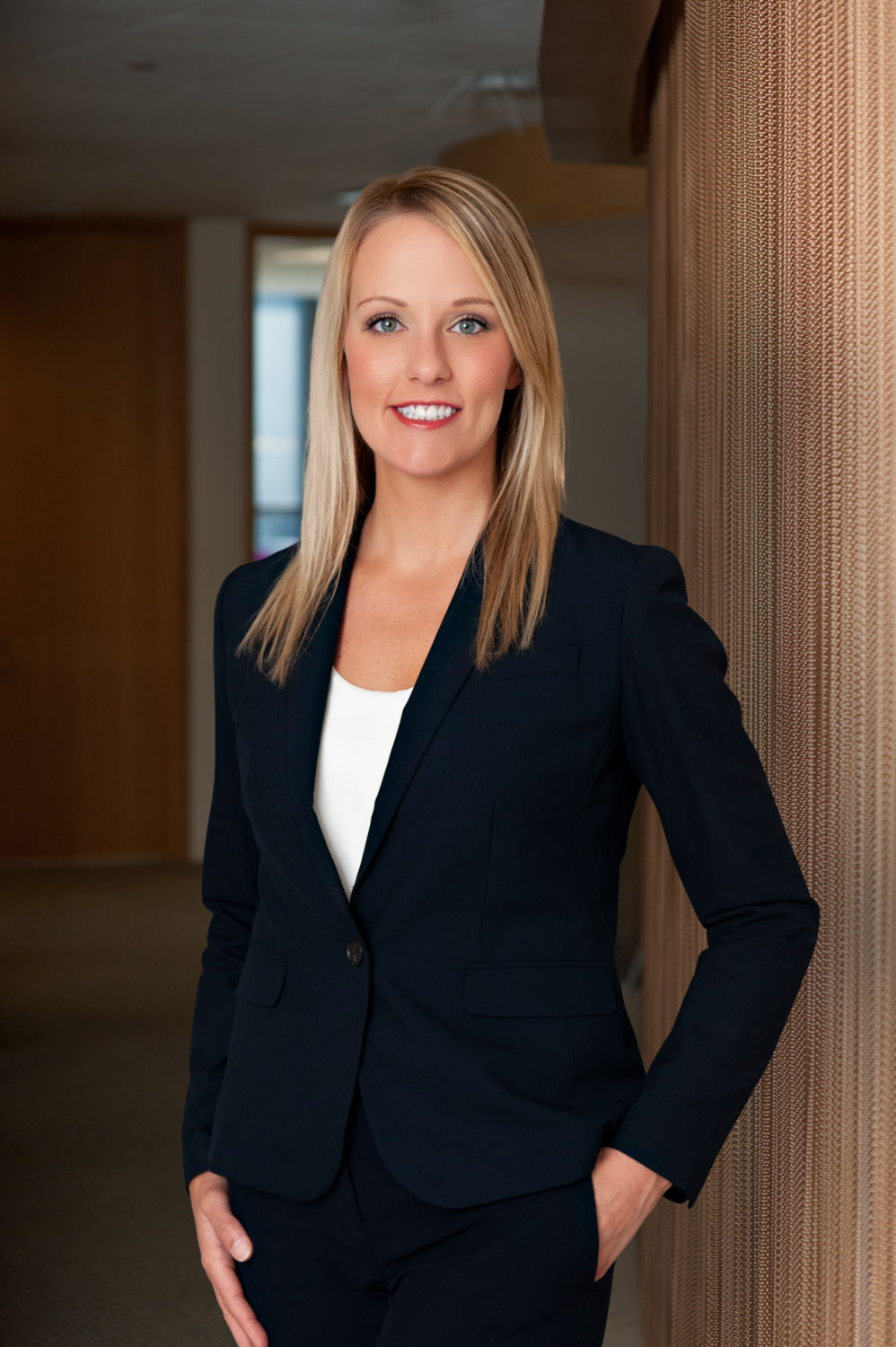 Professional portrait of Amanda Crain, attorney. She stands in a black suit in the hallway of a well lit office, meeting the camera with a confident gaze and smile. 