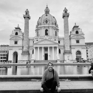 A woman sits in front of a white marble building with a proud dome in a black and white photograph. She leans toward the camera, smiling. 