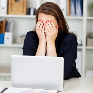 A photo of a woman sitting in front of a laptop in a black suit jacket. She's holding her hands over her face as she sits in a brightly lit office with bookshelves in the background.  