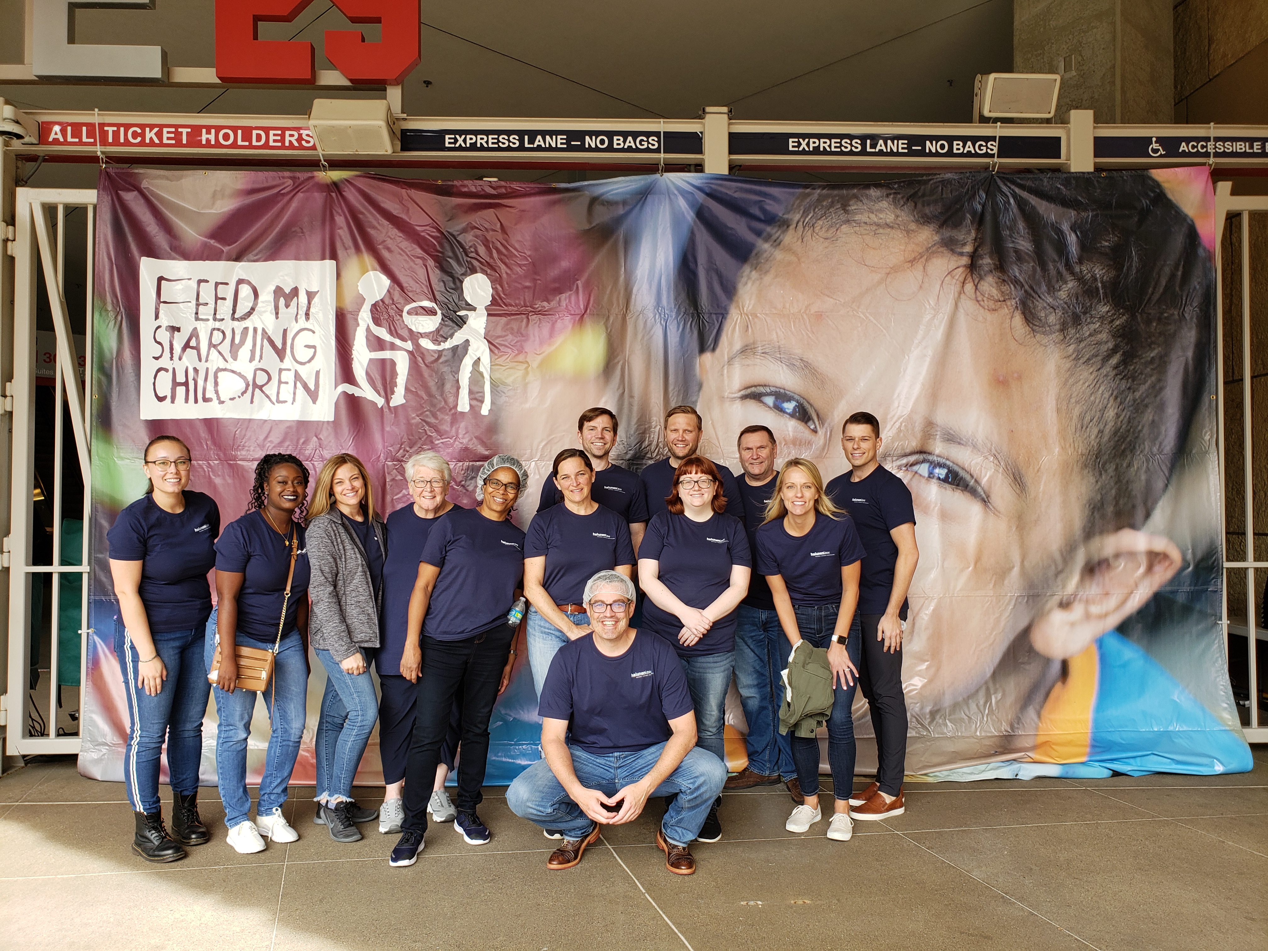the Halunen Law team stand in front of a colorful banner reading "Feed My Starving Children" while wearing matching dark blue t-shirts. Smiling, the team greets the camera, ready for the FMSC event.