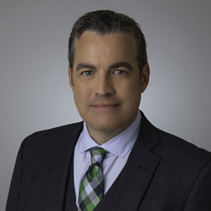 Chris Moreland, attorney, sits for a portrait in a studio setting. Wearing a professional gray suit, the man meets the camera with a serious gaze. 