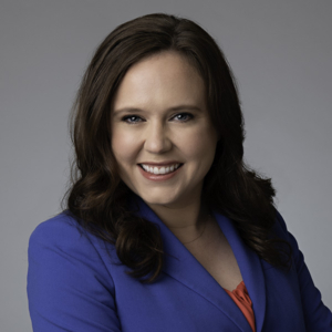 A close-cropped photo of Emma Denny. She wears a bright blue suit and smiles at the camera from a studio setting.