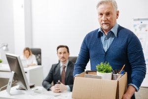 An older man in an office setting holds a box filled with items from his desk as a younger man looks on.