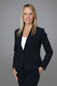 Attorney, Amanda Crain. stands for a portrait in a studio setting. She wears her blond hair down and a sleek black suit. She stands with one hand in her pocket, smiling confidently at the camera.
