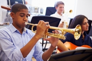 A young man plays the trumpet in a band classroom setting as a girl plays the guitar beside him and another boy holds an instrument in the background. 