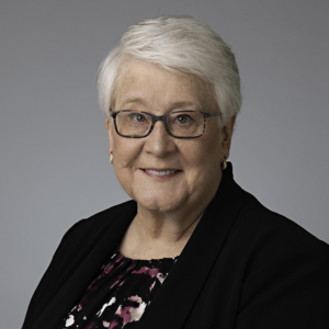A studio portrait of attorney, Susan Coler. She has short white hair, glasses, and wears a black suit. 
