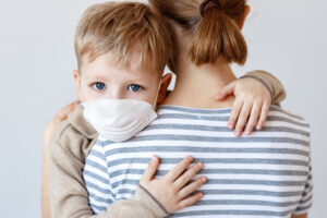 A little boy clings to his mother while wearing a medical mask. He faces the camera while his mother faces away.